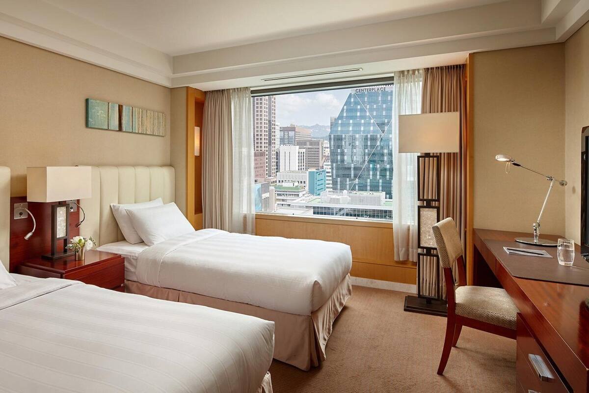 A hotel room with two beds, a desk, a chair, a lamp, and a view of a cityscape.