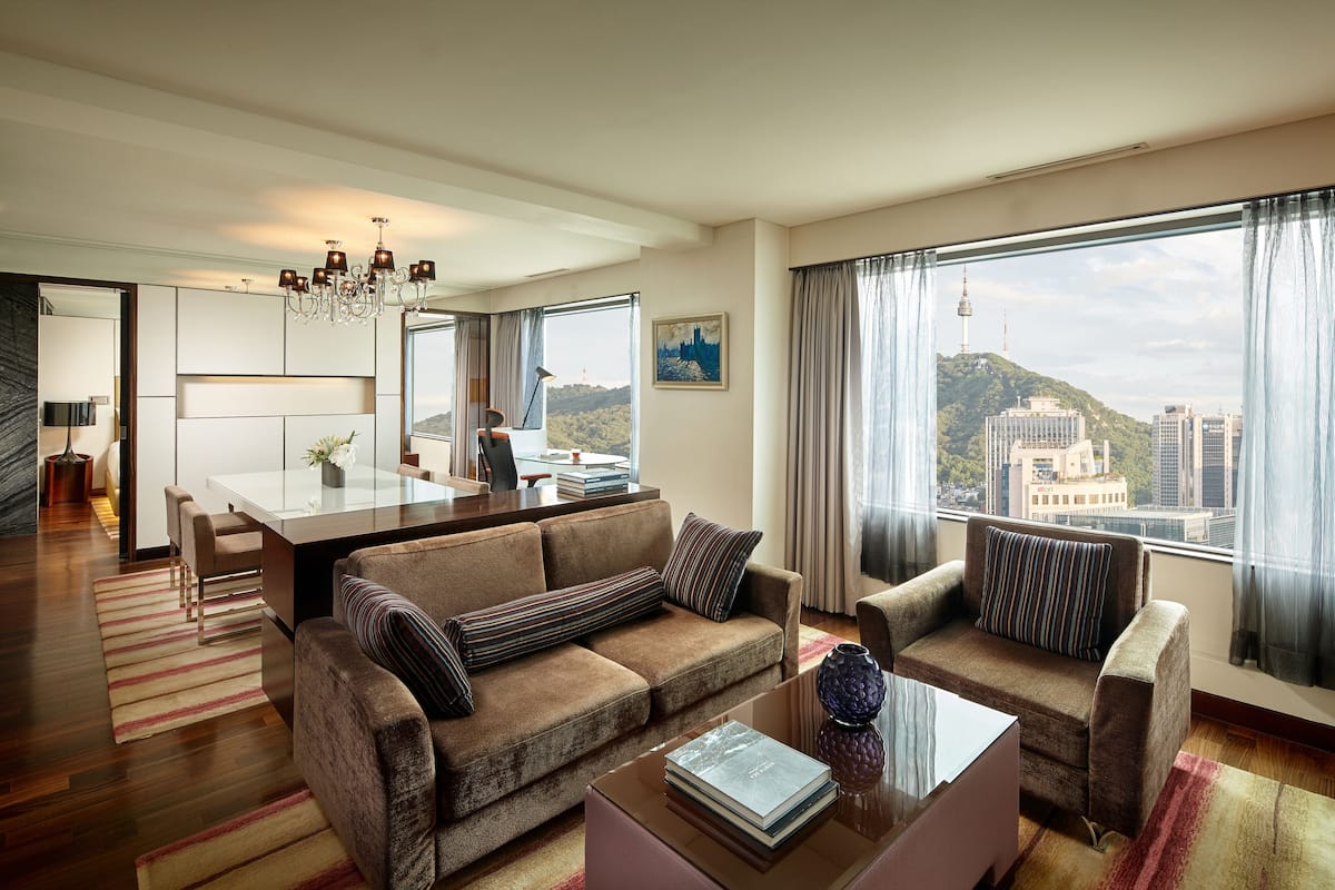 A modern hotel room with a sofa, armchair, dining table, and a view of the cityscape.