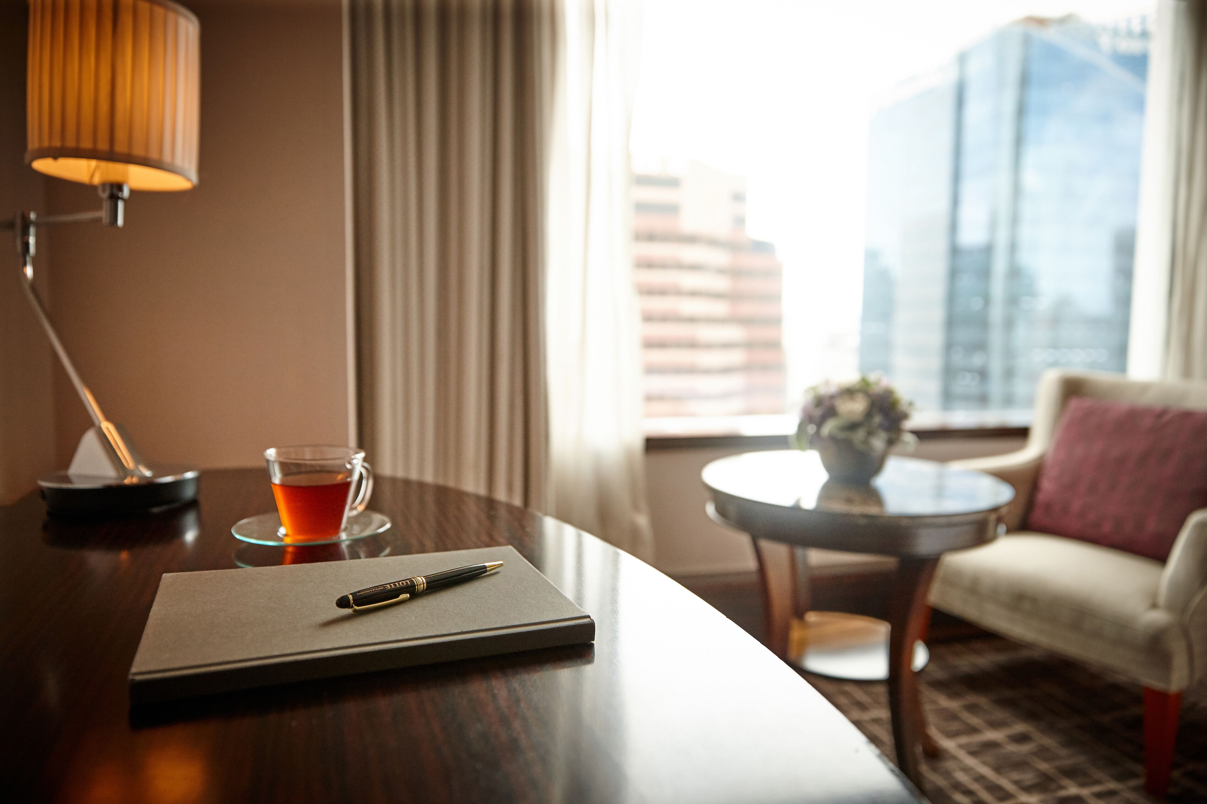 Habitación de hotel con un escritorio de madera, una lámpara, una taza de té, un cuaderno y un bolígrafo. Hay una silla y una mesita con un florero.