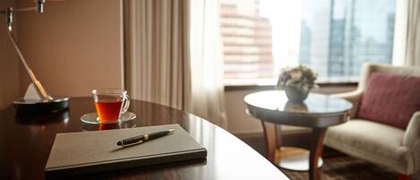Habitación de hotel con un escritorio de madera, una lámpara, una taza de té, un cuaderno y un bolígrafo. Hay una silla y una mesita con un florero.