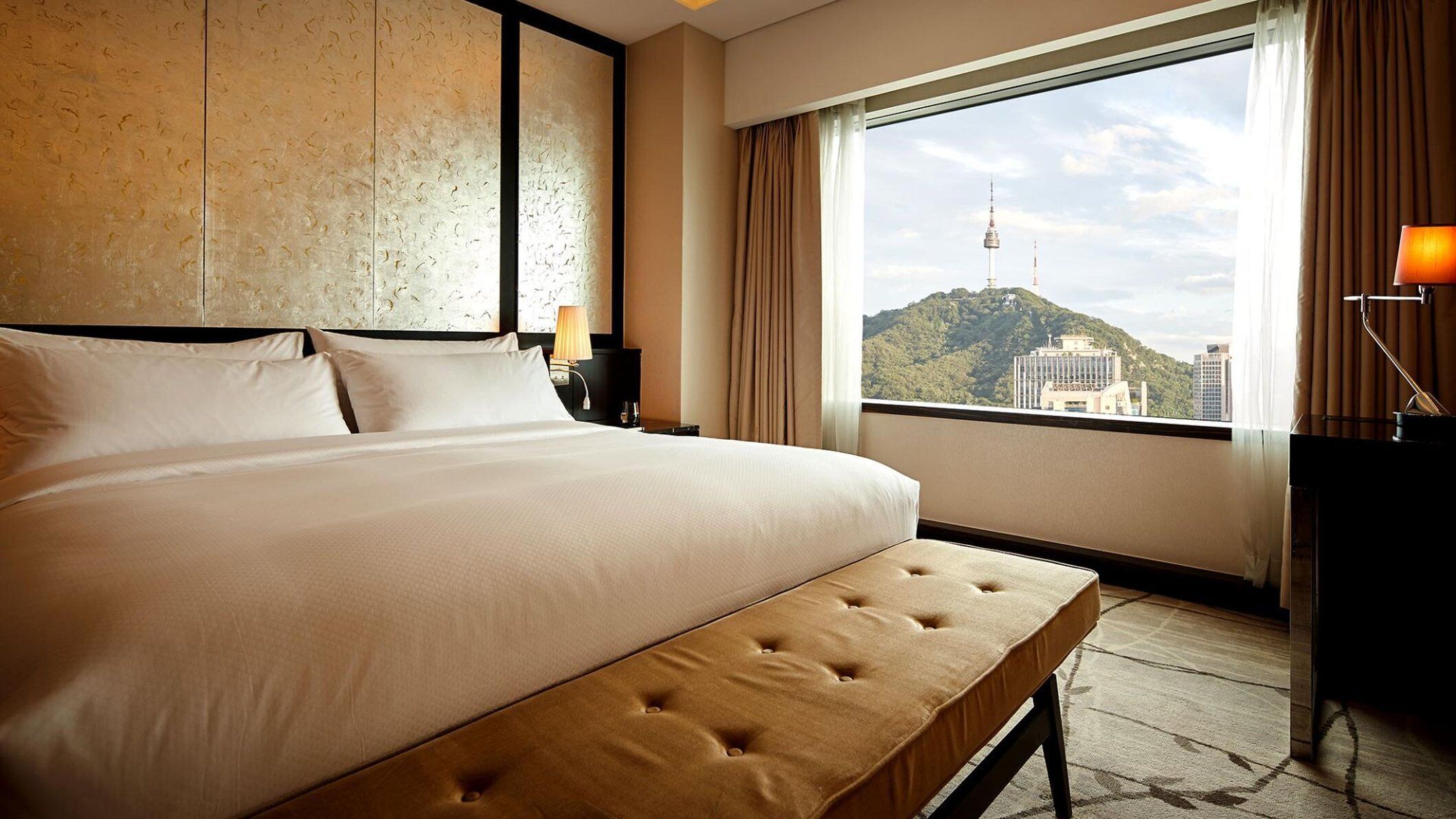 A hotel room with a large bed, a bench, a nightstand, a lamp, and a view of a cityscape through a large window.