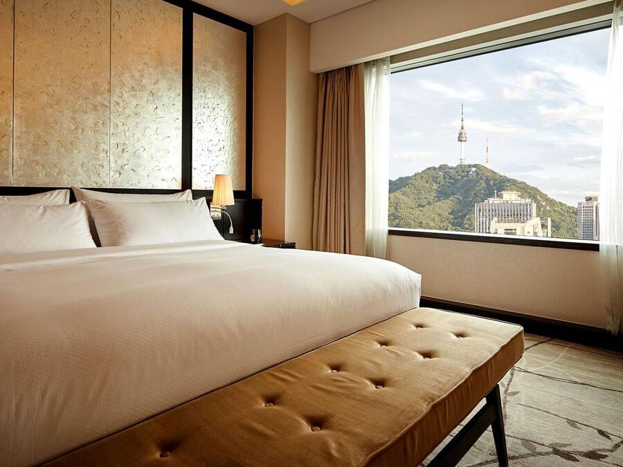 A hotel room with a large bed, a bench, a nightstand, a lamp, and a view of a cityscape through a large window.