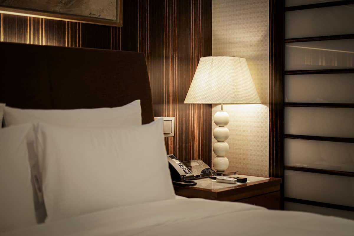 A neatly made bed with white linens, a wooden headboard, a bedside lamp, a telephone, and a wall-mounted light switch.