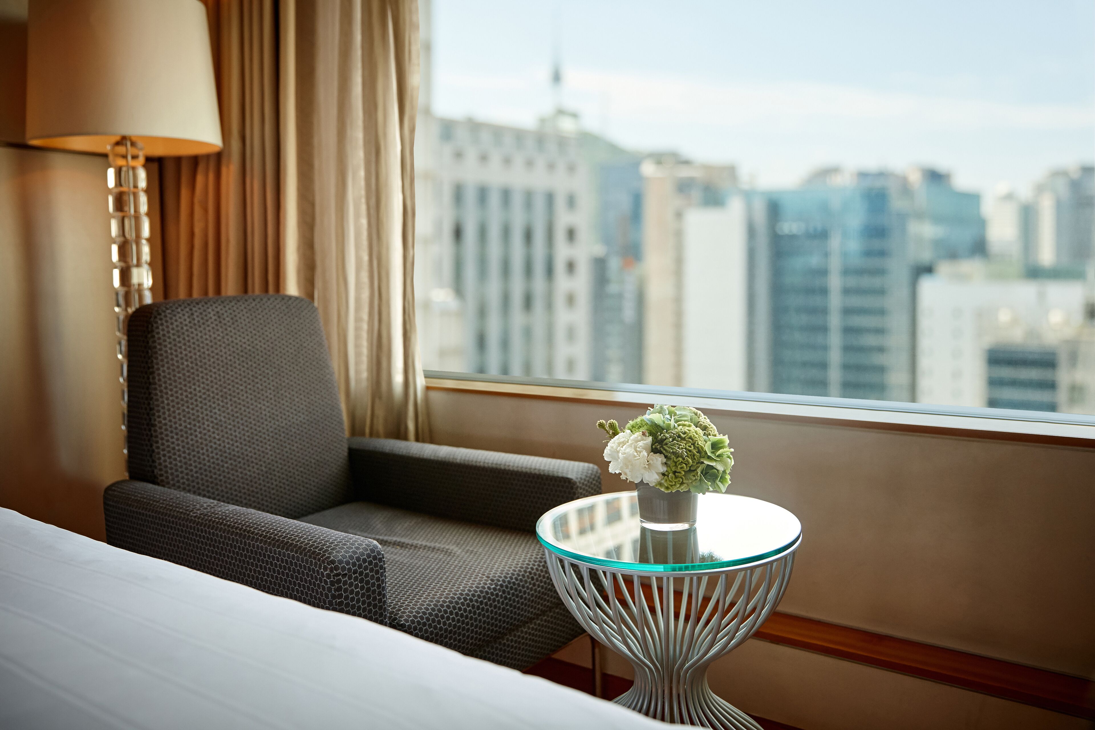 Een hotelkamer met een bed, een stoel, een tafeltje met een bloevas, een lamp en een uitzicht op een stadsgezicht door een raam.
