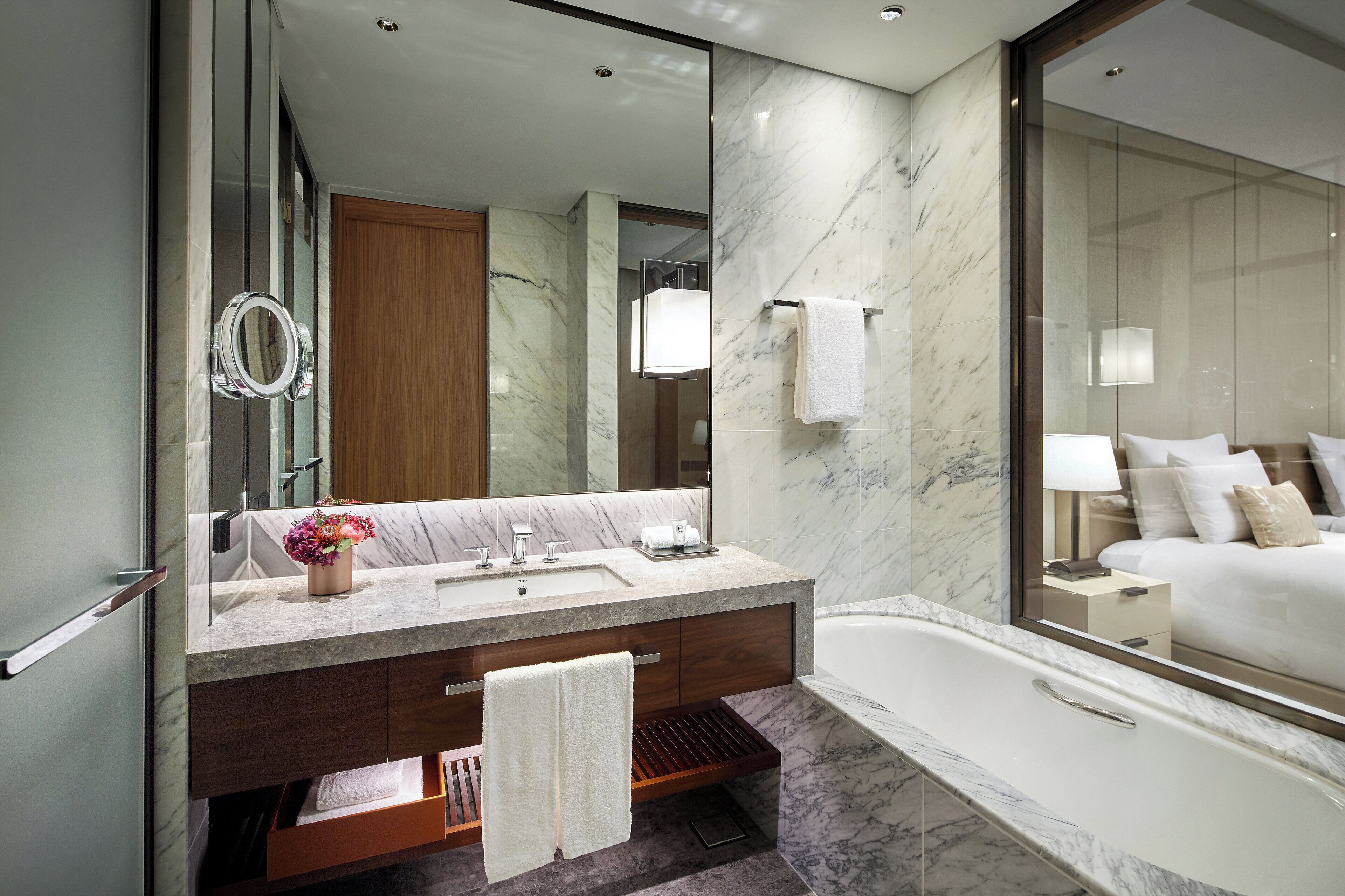 A modern bathroom with a marble sink, a large mirror, a wooden vanity, a bathtub, and a nightstand with a lamp.