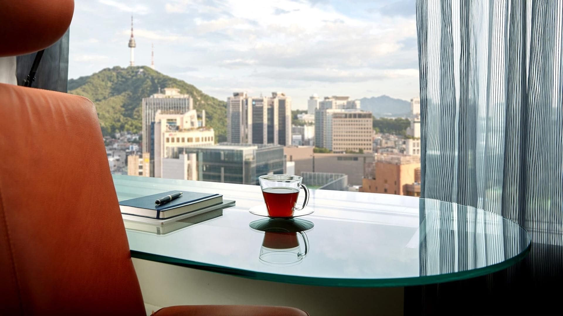 Una habitación moderna con una mesa de vidrio, un libro y una taza de té, con vistas a un paisaje urbano con edificios y una colina.