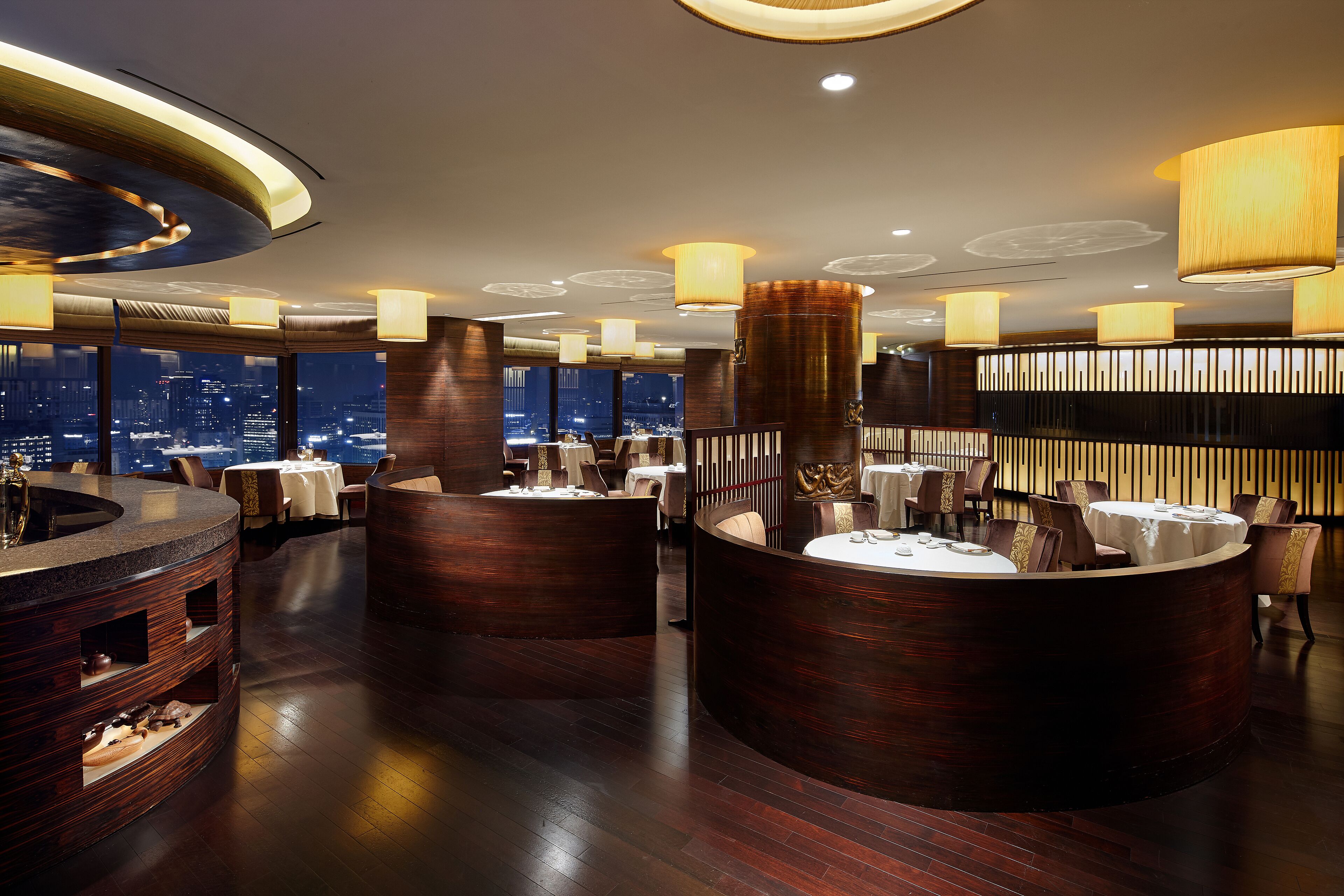 A modern restaurant with round tables and dark wood accents, featuring a city view.