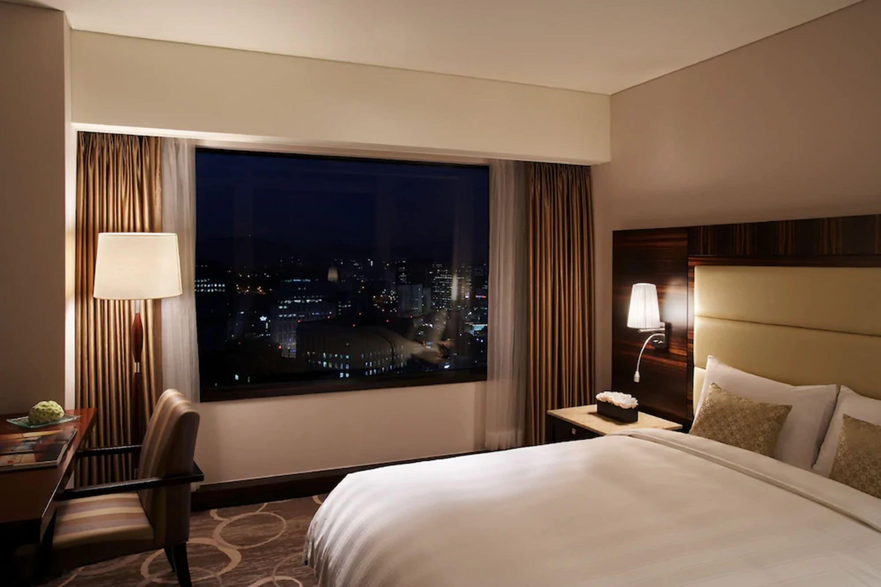 Ein Hotelzimmer mit großem Fenster, das einen Blick auf die beleuchtete Stadt bei Nacht bietet, einem Bett mit weißer Bettwäsche, einem Nachttisch mit Lampe und einem Sessel.