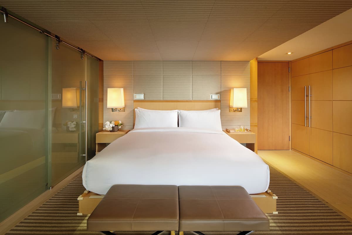 A modern hotel room with a large bed, bedside tables with lamps, a wooden wardrobe, and a glass partition.