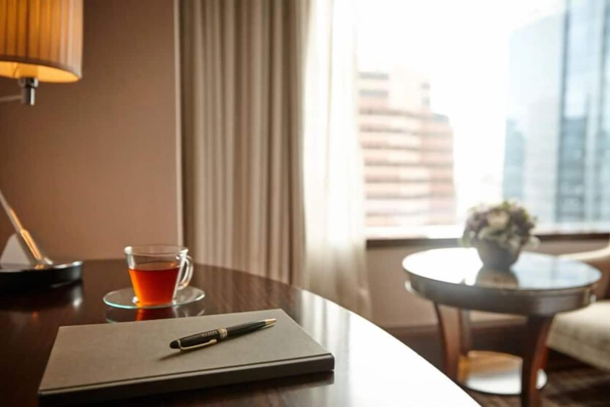 A hotel room with a desk, a cup of tea, a notebook, a pen, a lamp, a chair, and a view of the cityscape.