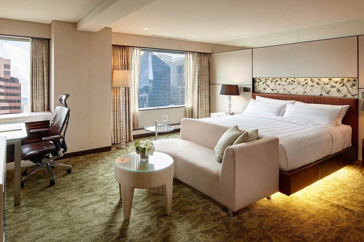 A hotel room with a bed, a sofa, a desk, a chair, a small table, and a view of a cityscape through large windows.