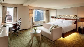 Habitación de hotel con cama, sofá, escritorio, silla, una pequeña mesa y vistas a la ciudad a través de amplias ventanas.