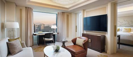A hotel room with a large window, a chandelier, a sofa, a chair, a TV, and a bed.