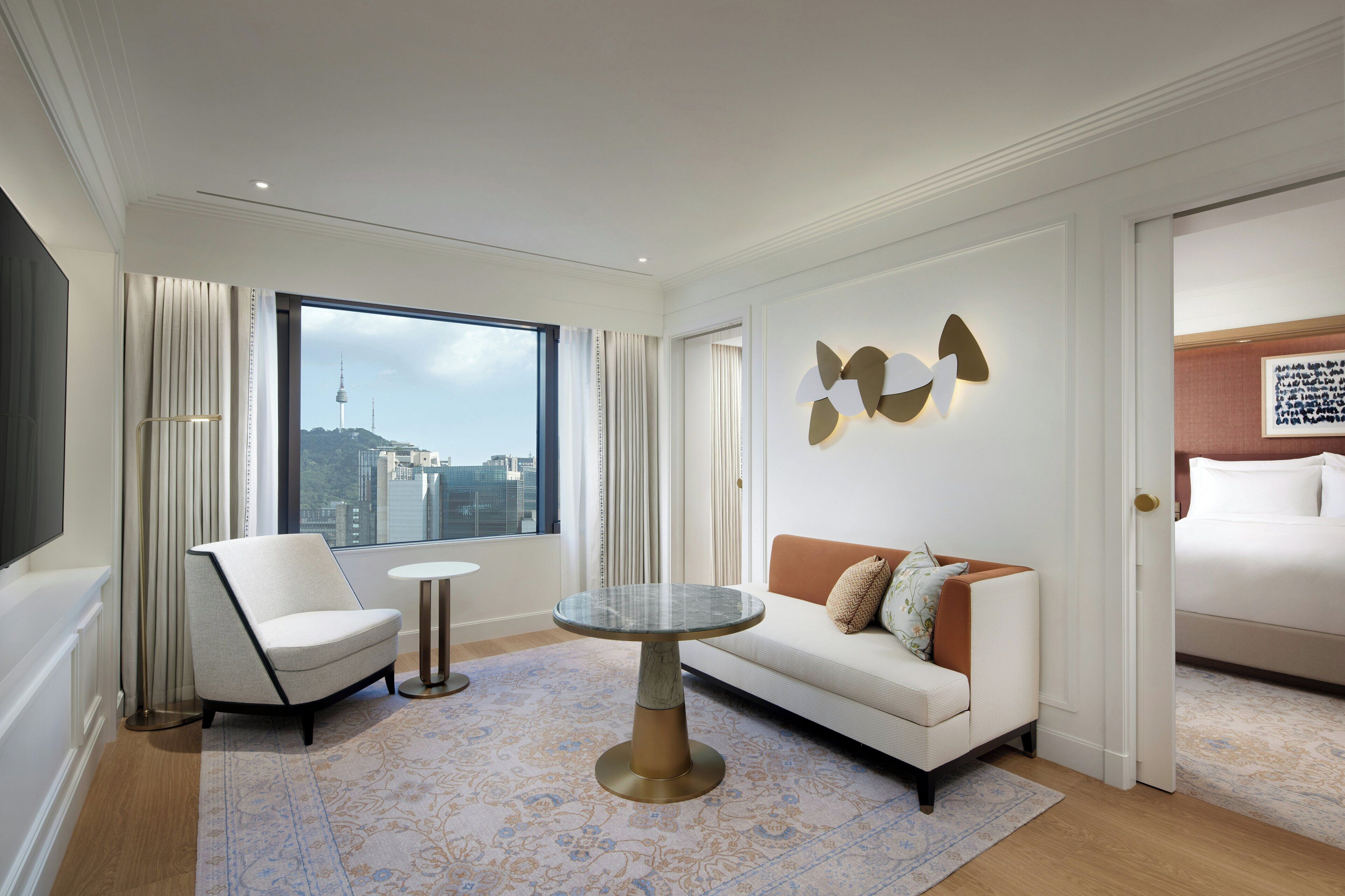A modern hotel room with a large window, a sofa, a chair, a small round table, and a decorative wall piece.