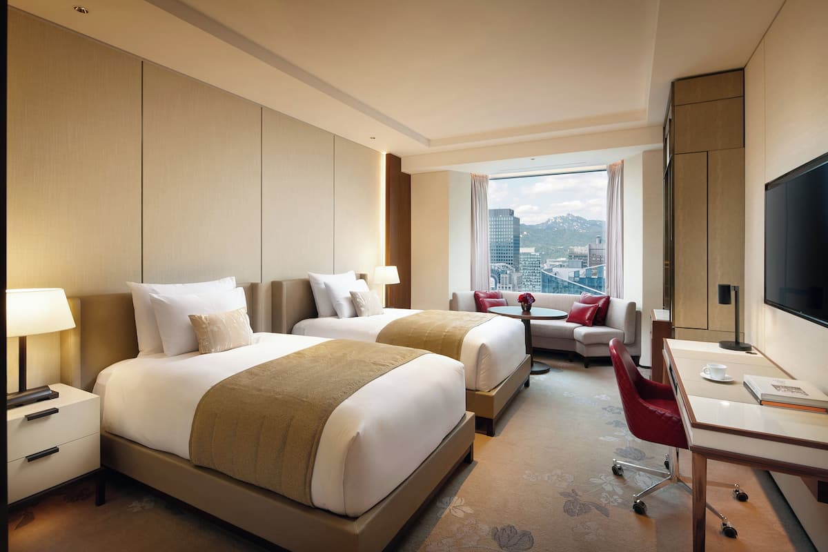 A hotel room with two beds, a desk, a chair, a TV, and a view of the city.