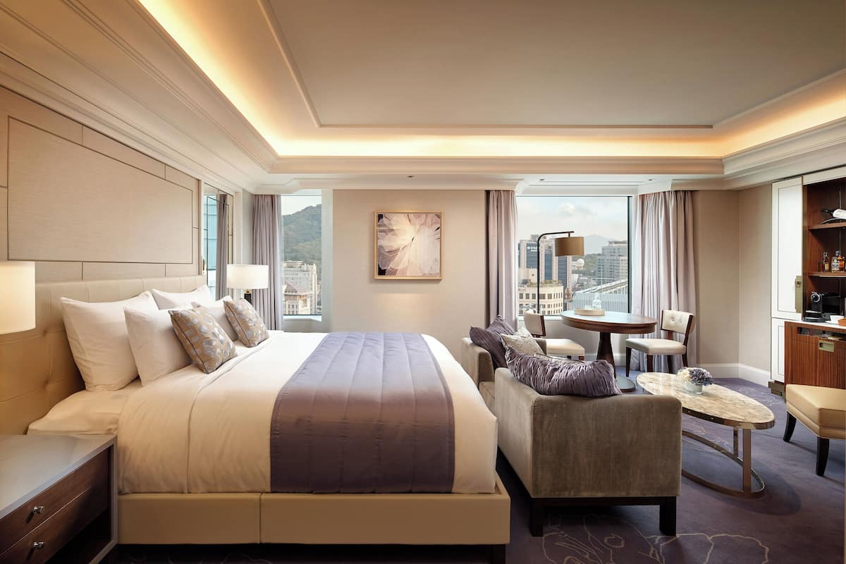 A modern hotel room with a large bed, a seating area with a table, and a view of the city.