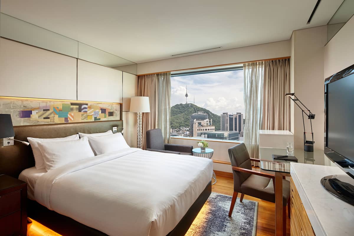 A hotel room with a large bed, a desk, a chair, a TV, and a view of a cityscape.