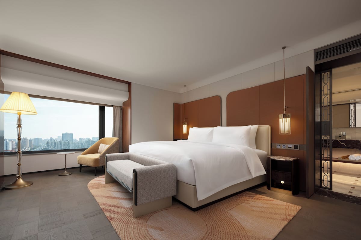 A modern hotel room with a large bed, a desk, a chair, a lamp, and a view of the city.