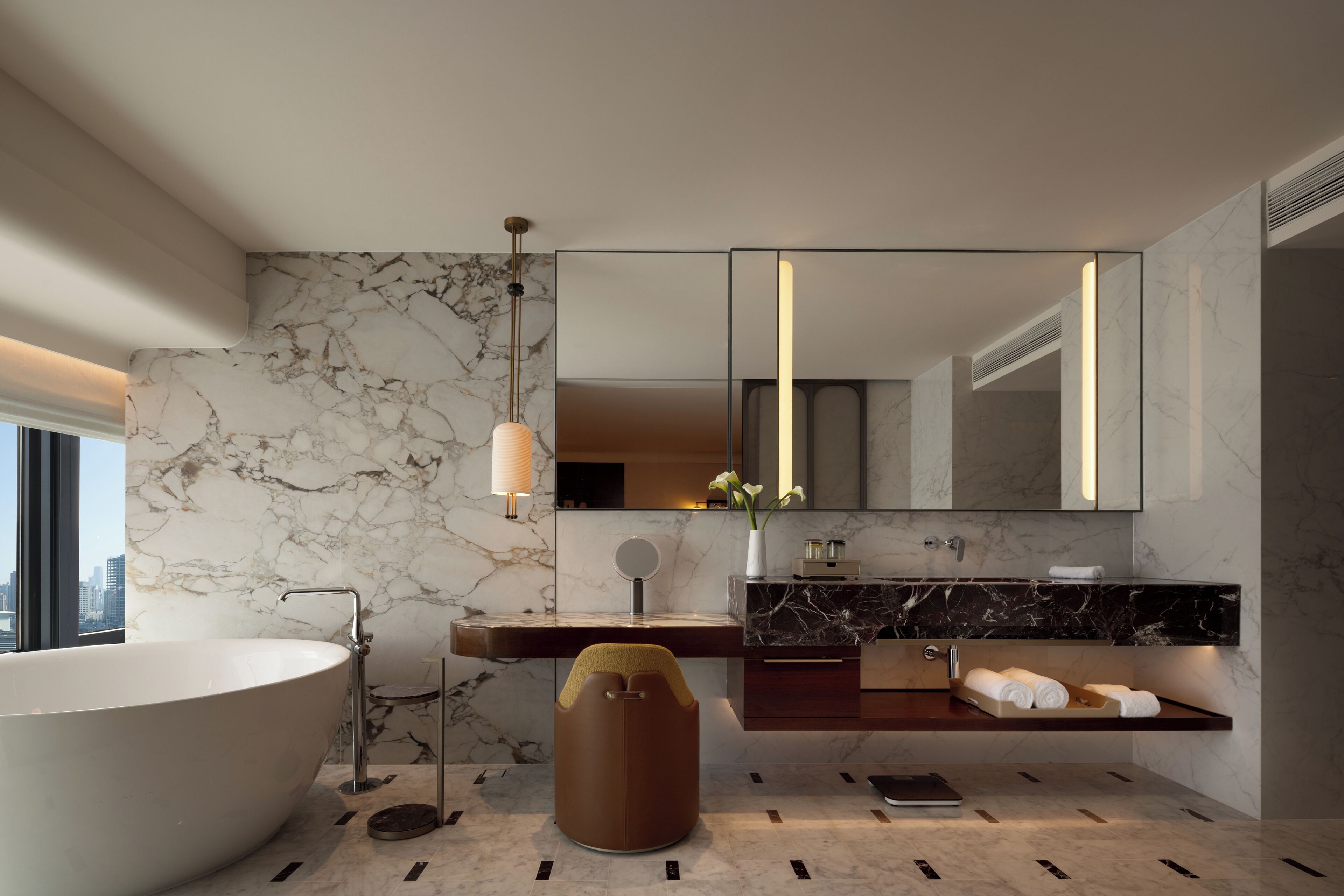 A modern bathroom with a freestanding bathtub, a vanity with a marble countertop, a large mirror, and a city view.