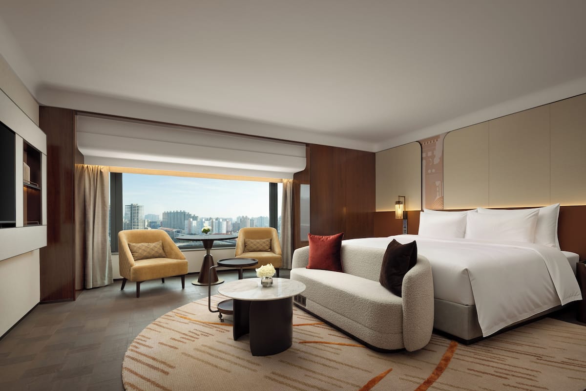 A modern hotel room with a large bed, a sofa, a small round table, two armchairs, a view of the city, and a large window.