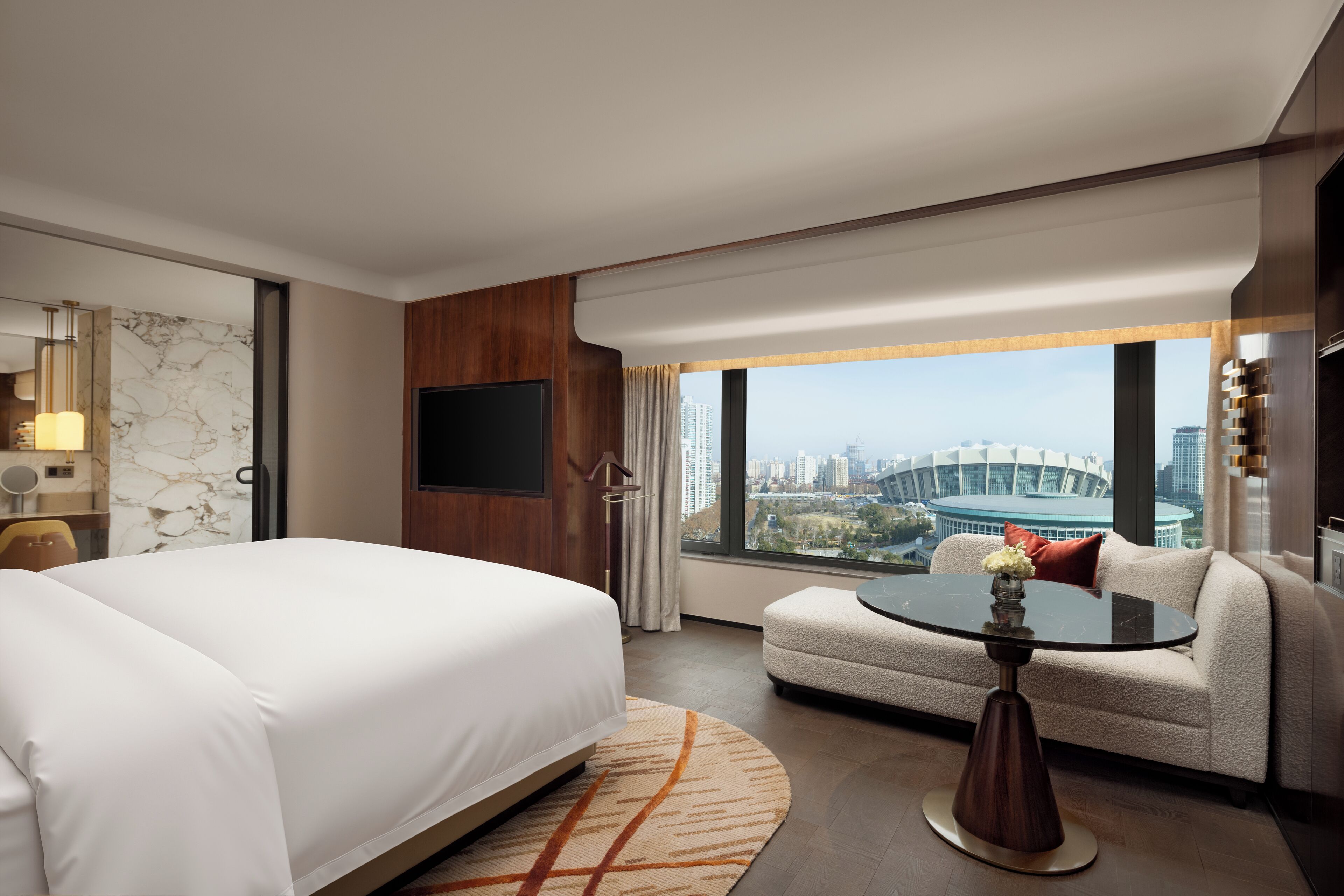 A hotel room with a bed, a sofa, a round table with a vase, a TV, and a large window with a city view.