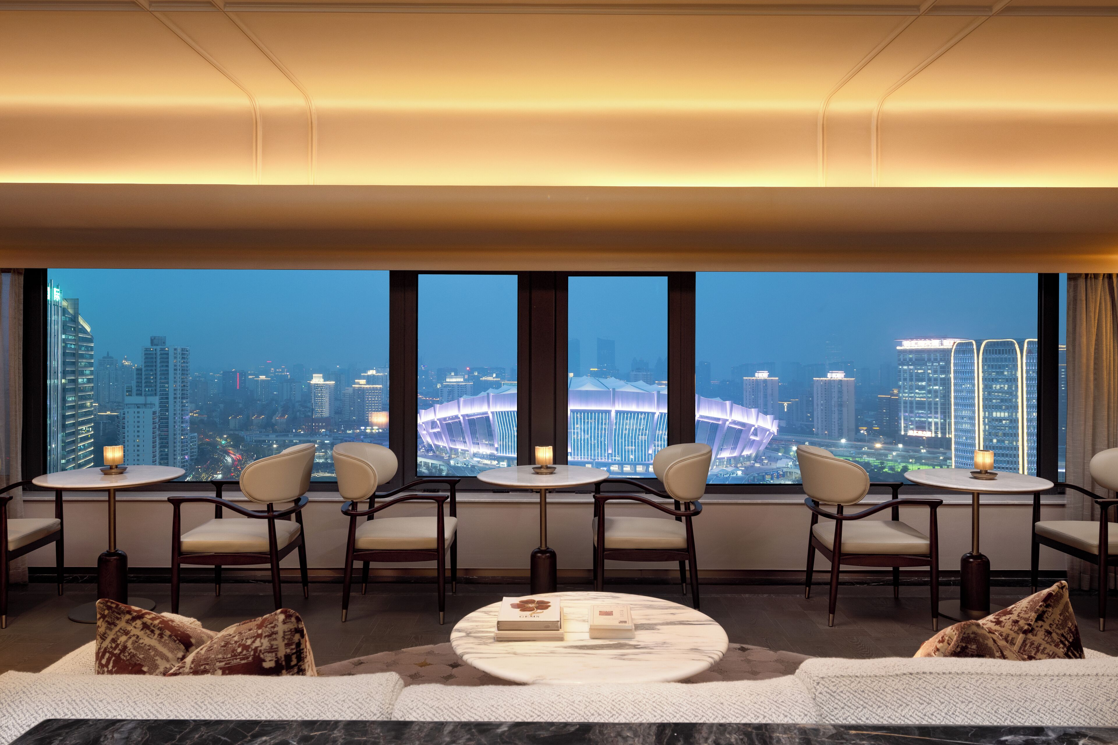 Habitación de hotel en un edificio alto con vista a la ciudad, una mesa redonda, cuatro sillas, un sofá y un ventanal grande.