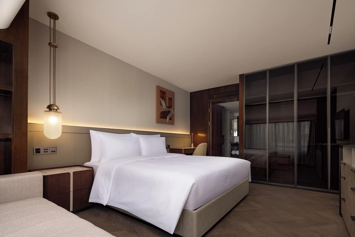 A modern hotel room with a large bed, bedside tables, a desk, a chair, a TV, and a sliding glass door to a balcony.