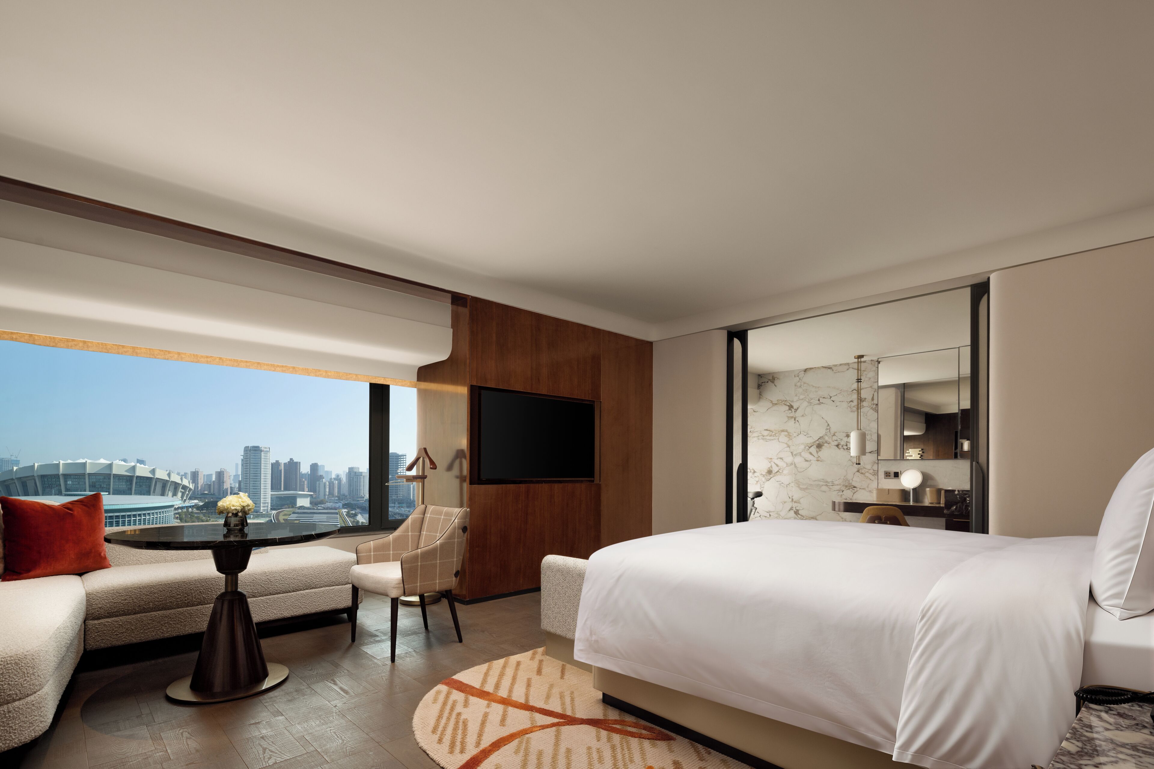 Quarto de hotel moderno com uma cama grande, área de estar com uma mesa, televisão na parede e vista para a cidade.