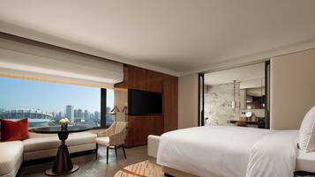 Quarto de hotel moderno com uma cama grande, área de estar com uma mesa, televisão na parede e vista para a cidade.