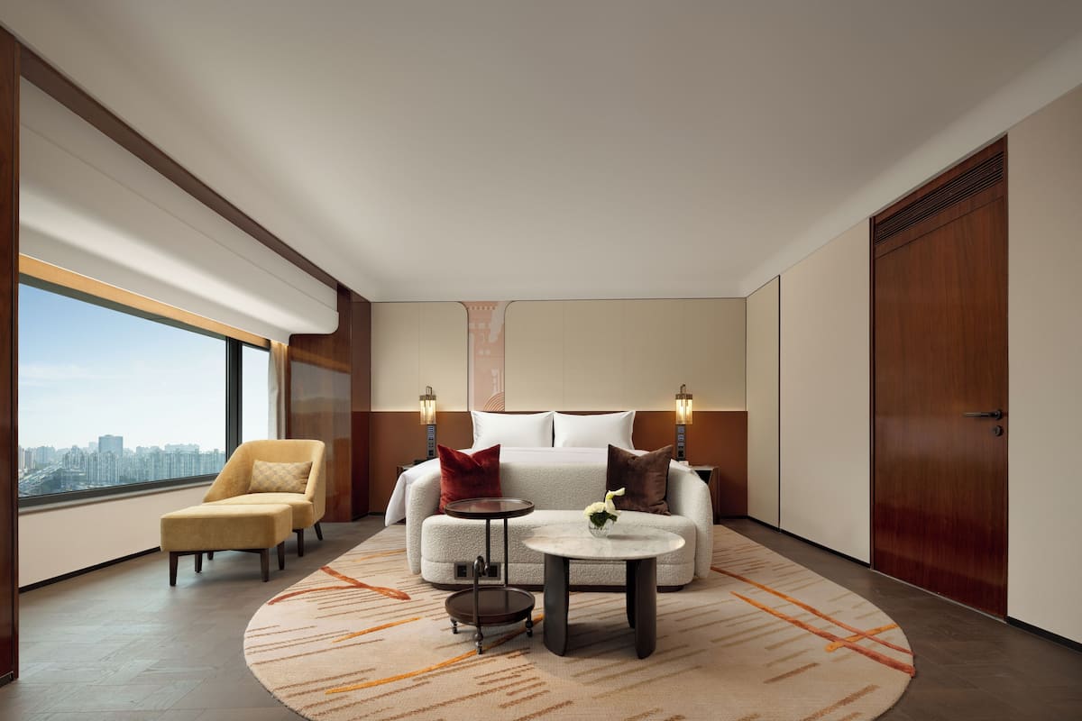 A hotel room with a large window, a sofa, a chair, a round table, and a bed.