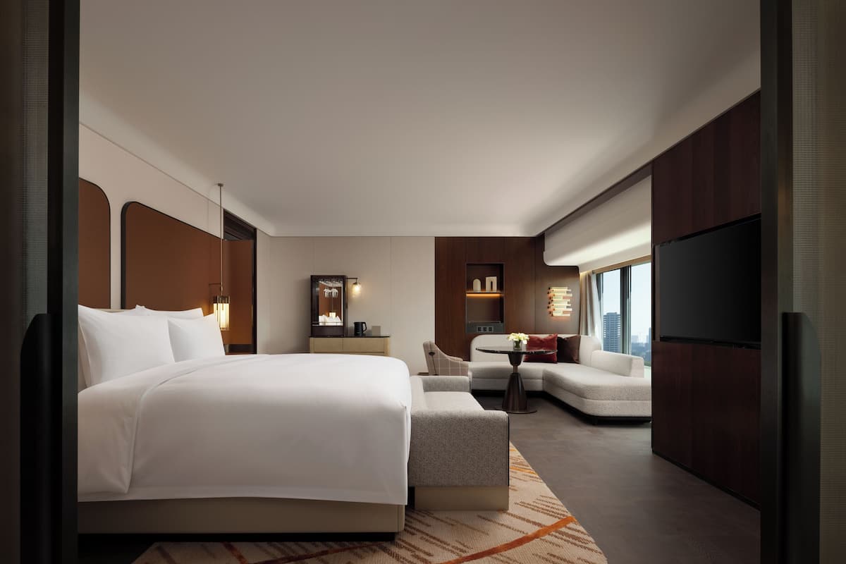 A modern hotel room with a large bed, a sofa, a small table, a TV, and a city view.