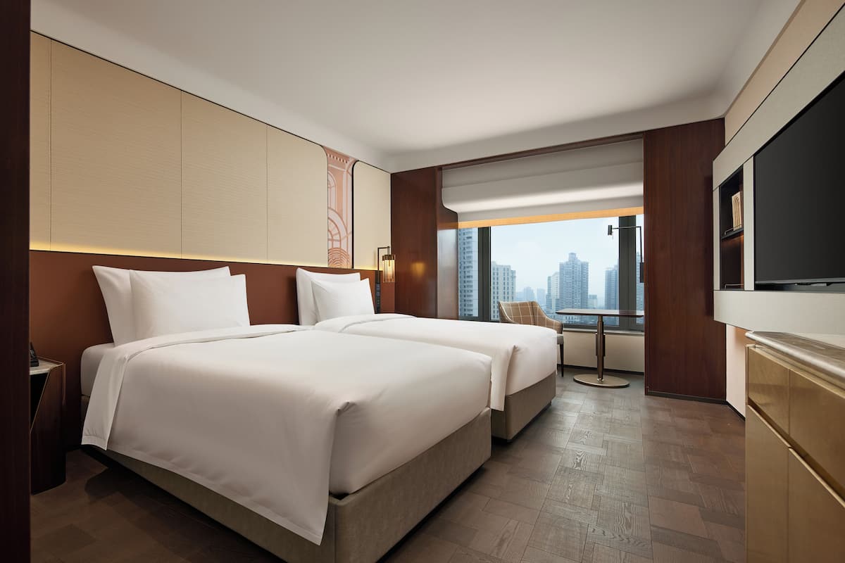 A hotel room with two beds, a TV, a desk, and a view of the city.