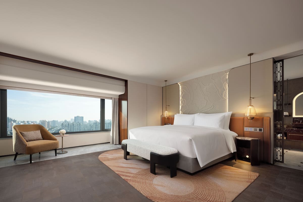 A modern hotel room with a large bed, a sitting area with a chair, a view of the city, and a bathroom with a vanity.