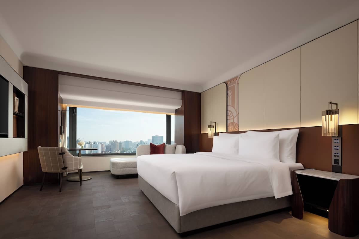 A modern hotel room with a large bed, a seating area with a chair, a side table, and a view of the city.