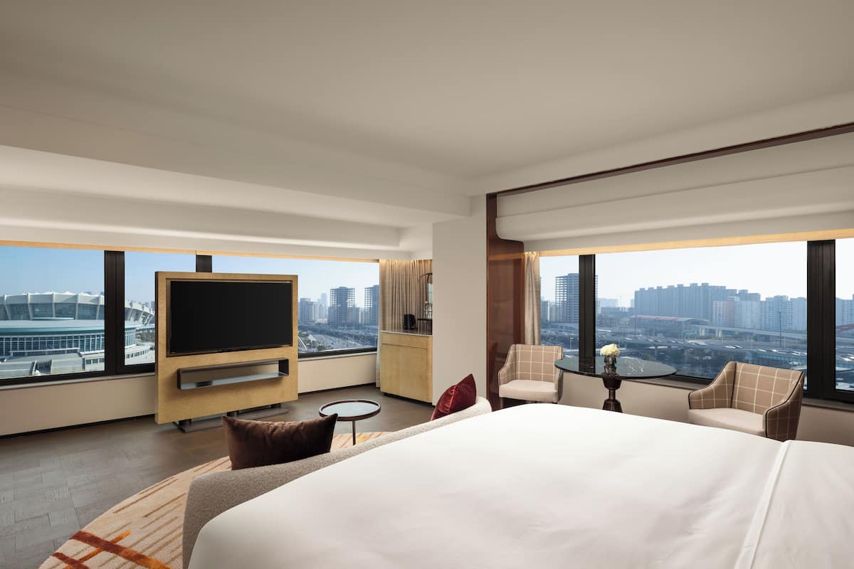 A modern hotel room with a large bed, a TV, a small table, a chair, and a view of the cityscape.