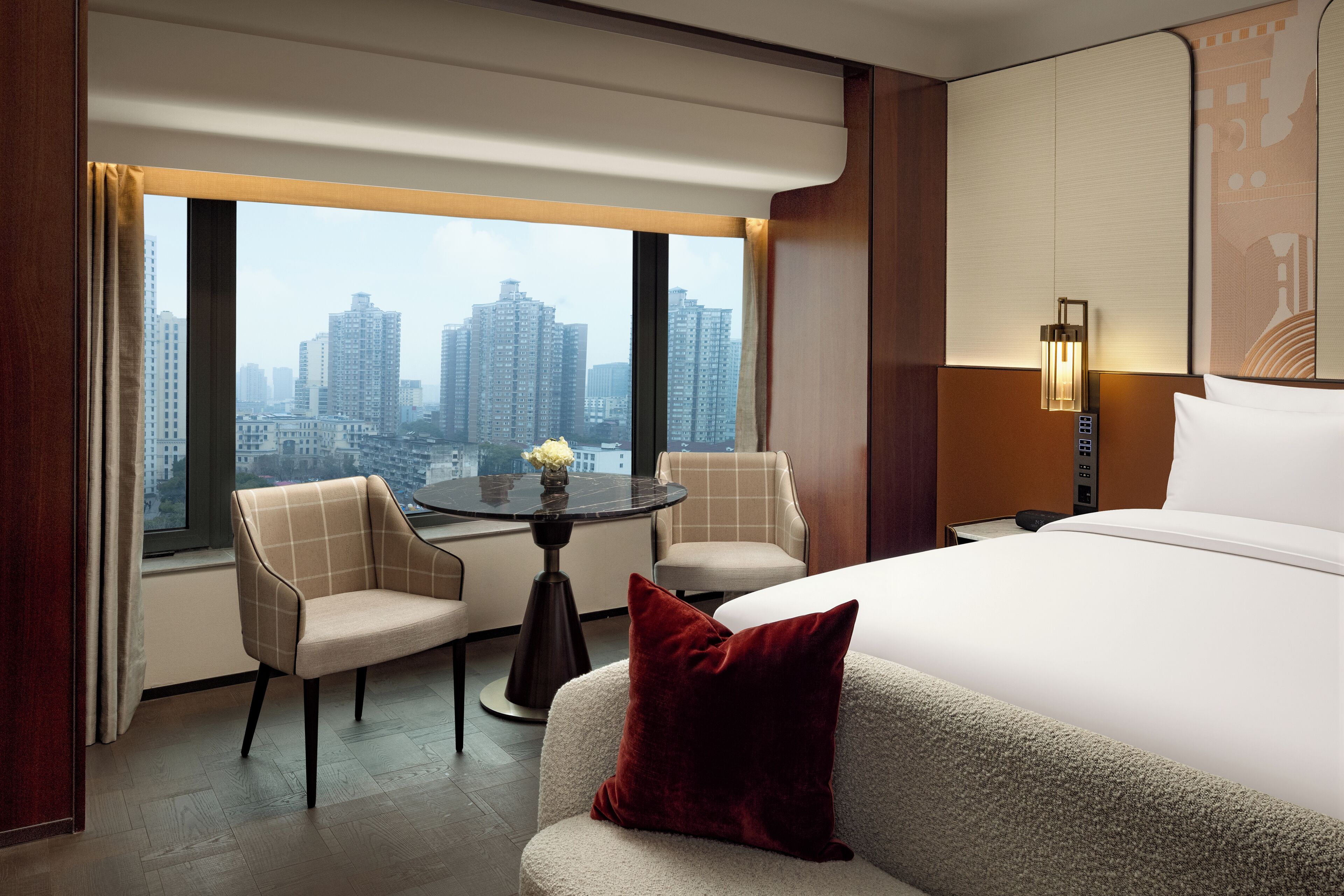Habitación de hotel con cama, dos sillones, una mesita con un jarrón, vista a la ciudad y una lámpara de pared.