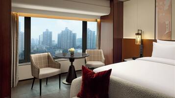 Habitación de hotel con cama, dos sillones, una mesita con un jarrón, vista a la ciudad y una lámpara de pared.