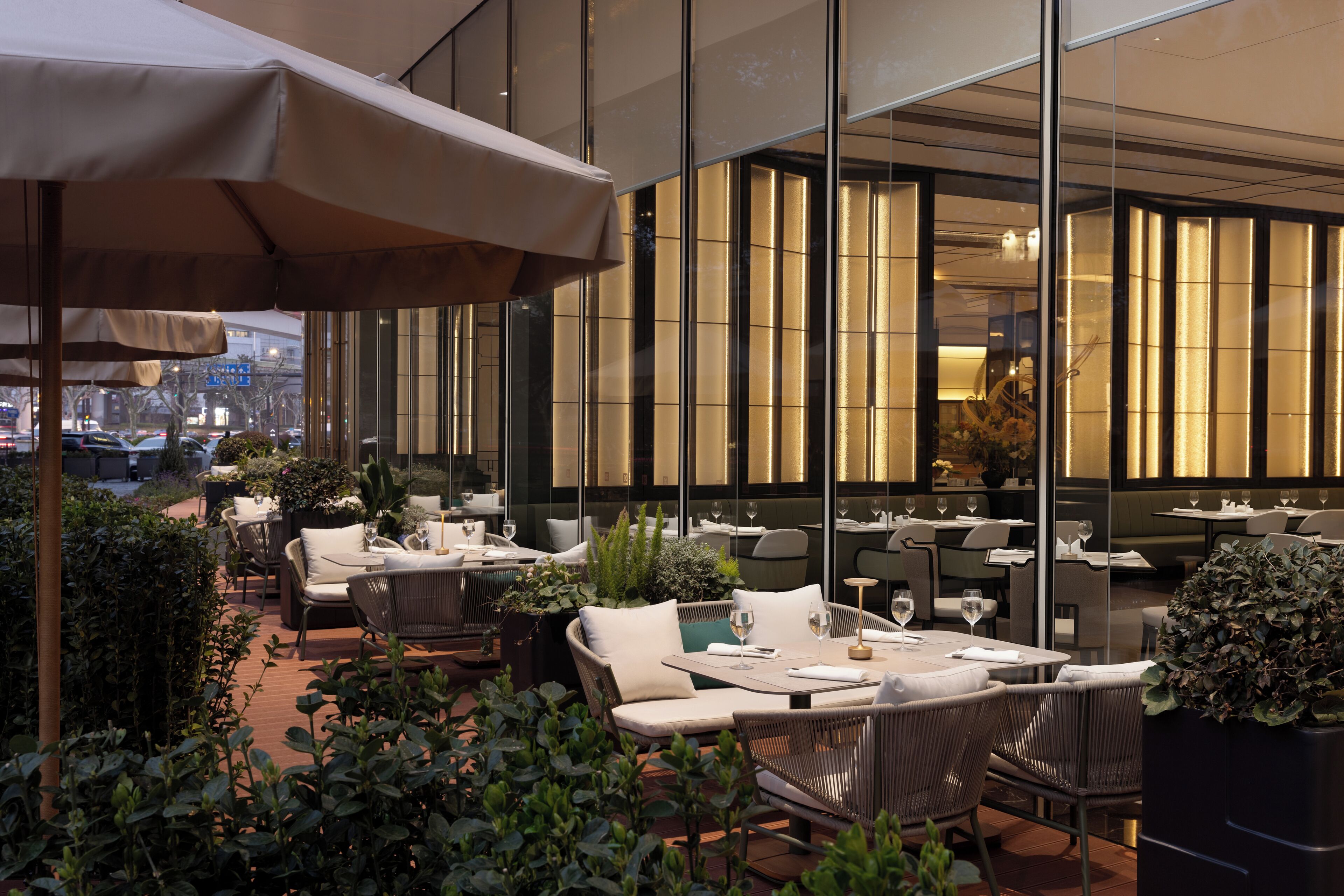An outdoor dining area with tables and chairs, surrounded by greenery and a glass wall.