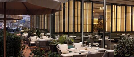 An outdoor dining area with tables and chairs, surrounded by greenery and a glass wall.