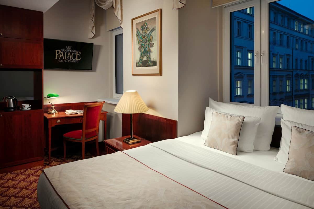 Een hotelkamer met een bed, een bureau, een stoel, een lamp, een lijst met kunst en uitzicht op een gebouw door het raam.