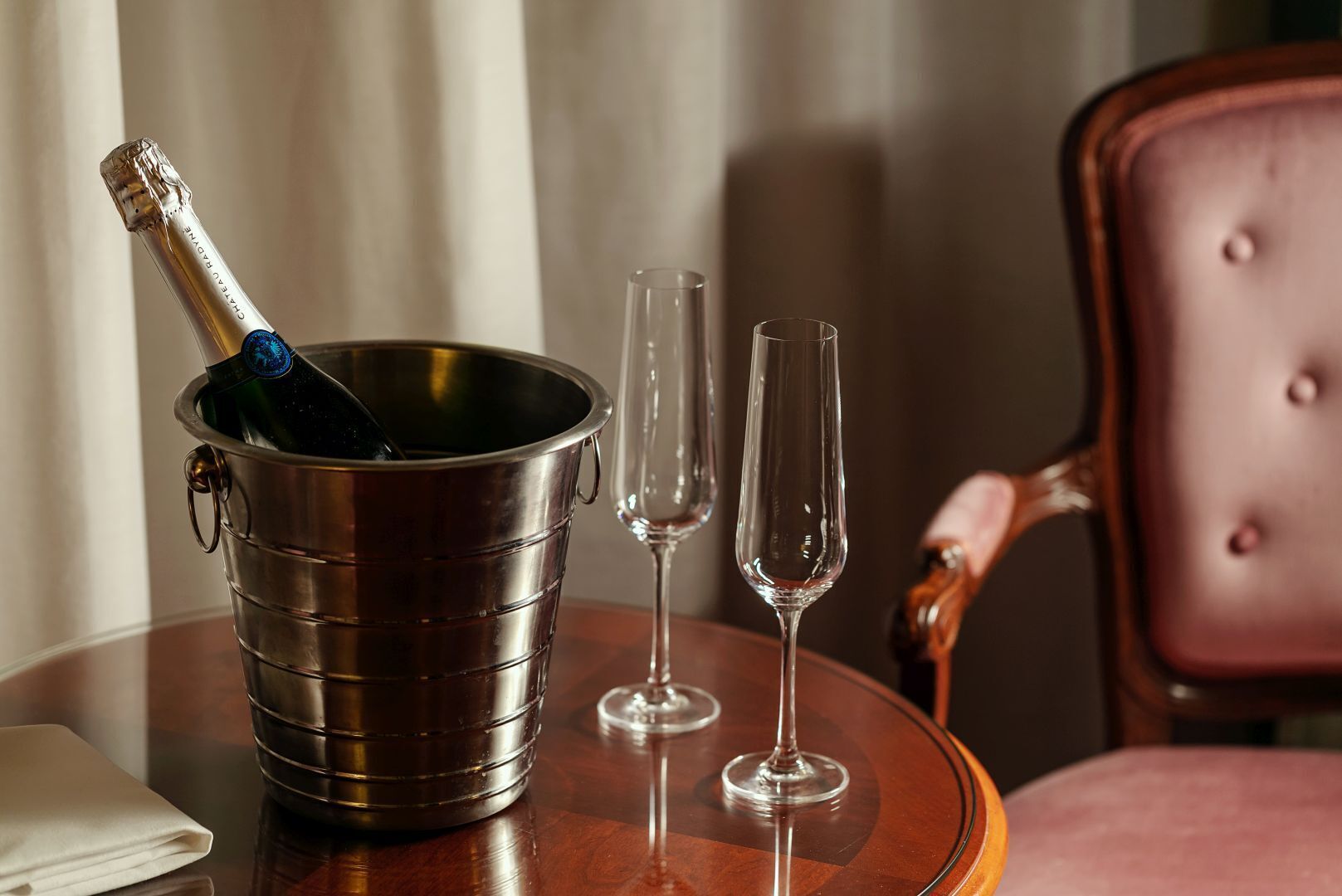 A metal bucket with a bottle of champagne, two empty wine glasses, and a folded napkin on a wooden table.