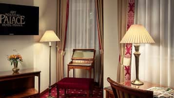 A hotel room with a wooden desk, a red upholstered bench, a standing lamp, a television displaying 'Art Nouveau Palace Hotel Prague', and a window with sheer curtains.