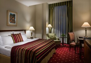 Quarto de hotel com uma cama grande, duas cadeiras, uma mesinha e uma escrivaninha.
