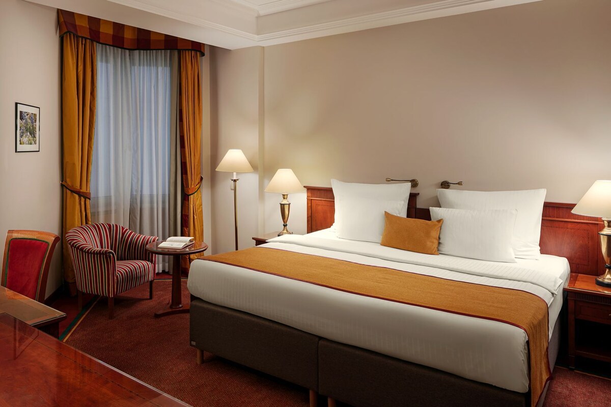 Una habitación de hotel con una cama grande, dos lámparas de noche, una silla, un escritorio y un ventanal con cortinas.