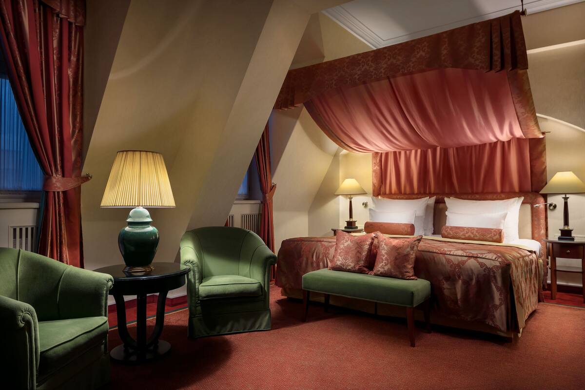 Een kamer met een hemelbed, twee lampen, een groene fauteuil, een groene bank en een kleine tafel.
