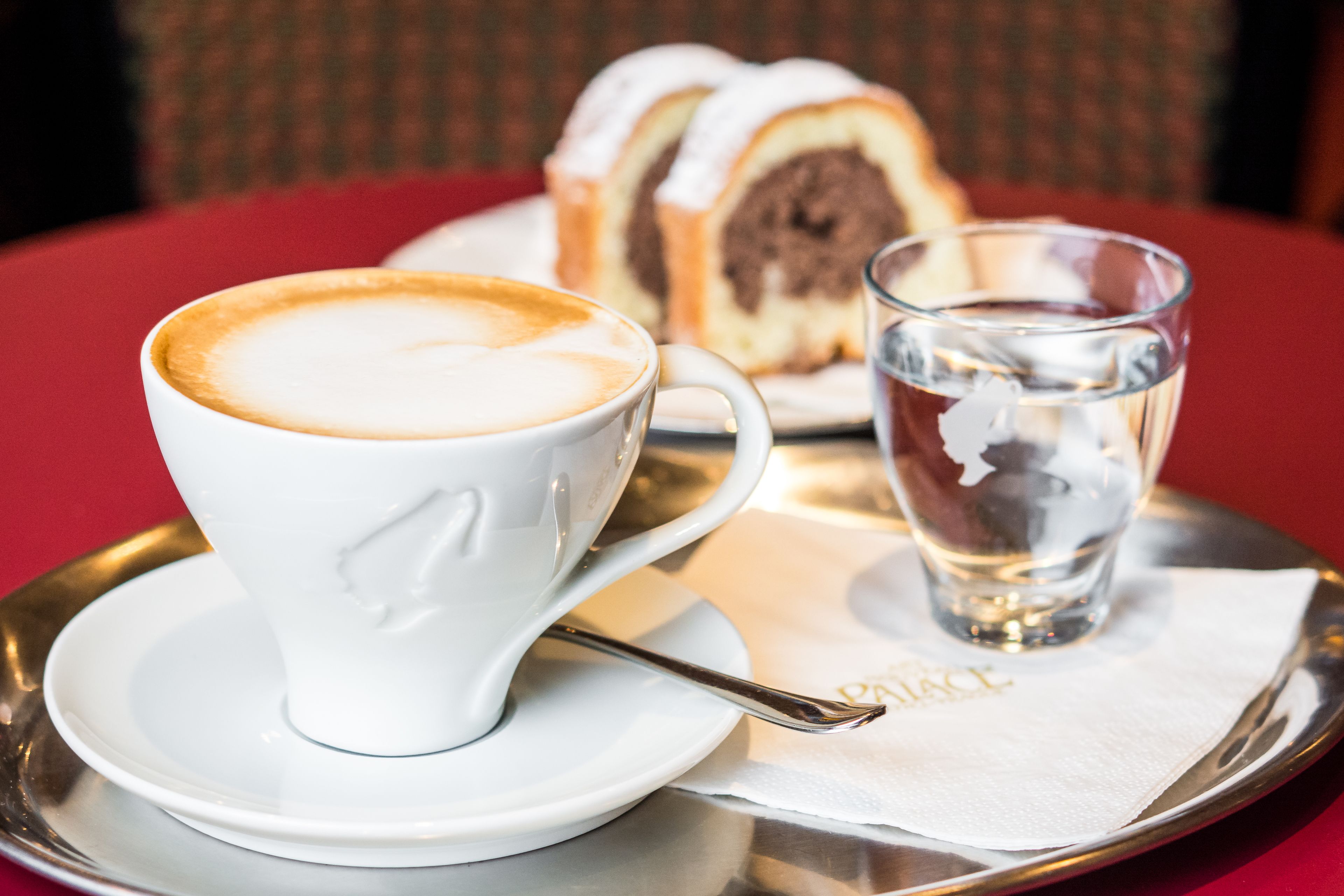 Una taza blanca de café con una cuchara sobre una bandeja, una porción de pastel y un vaso de agua.