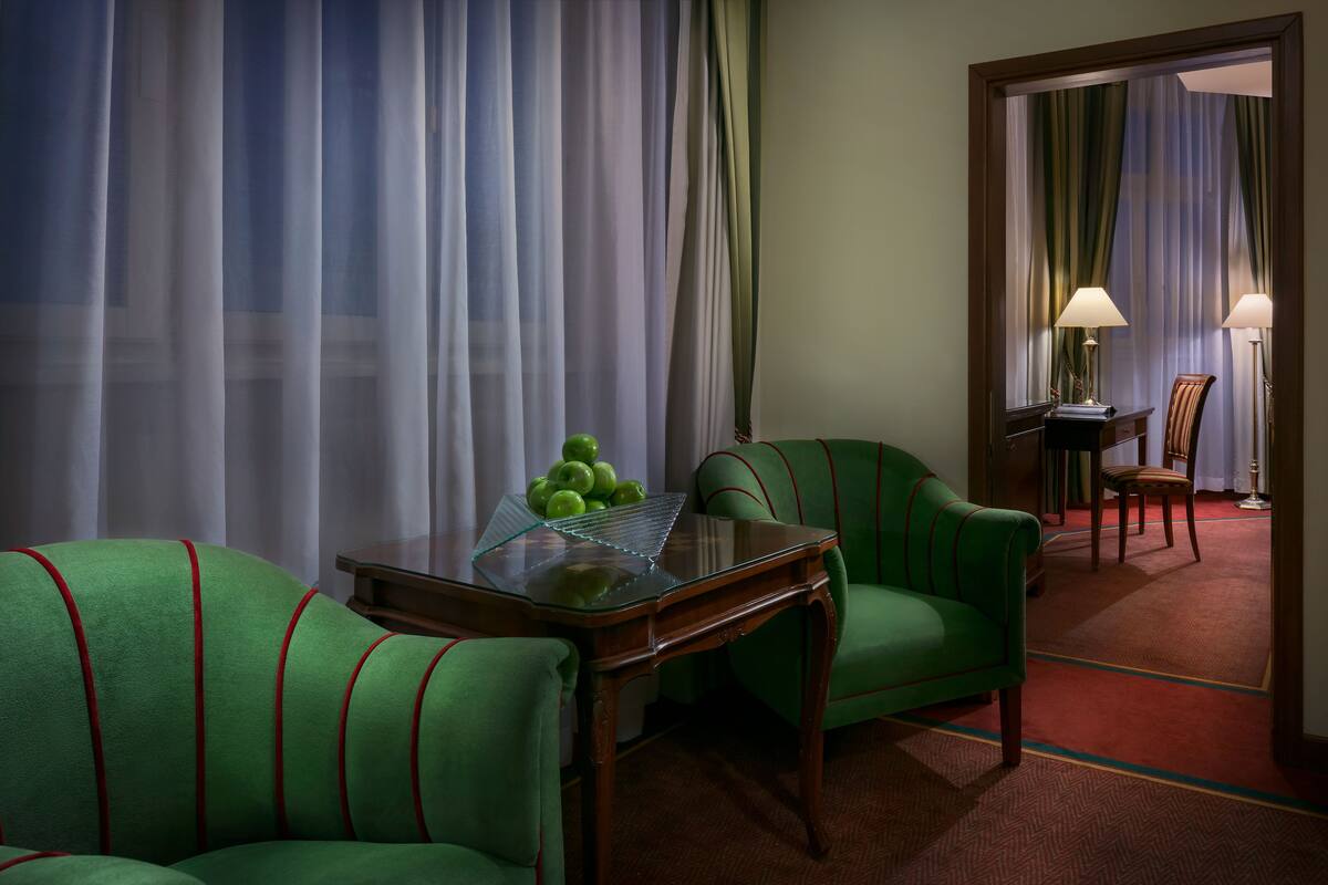 Een hotelkamer met een groene bank, een tafel met een glazen blad met appels, en uitzicht door een raam met dunne gordijnen.