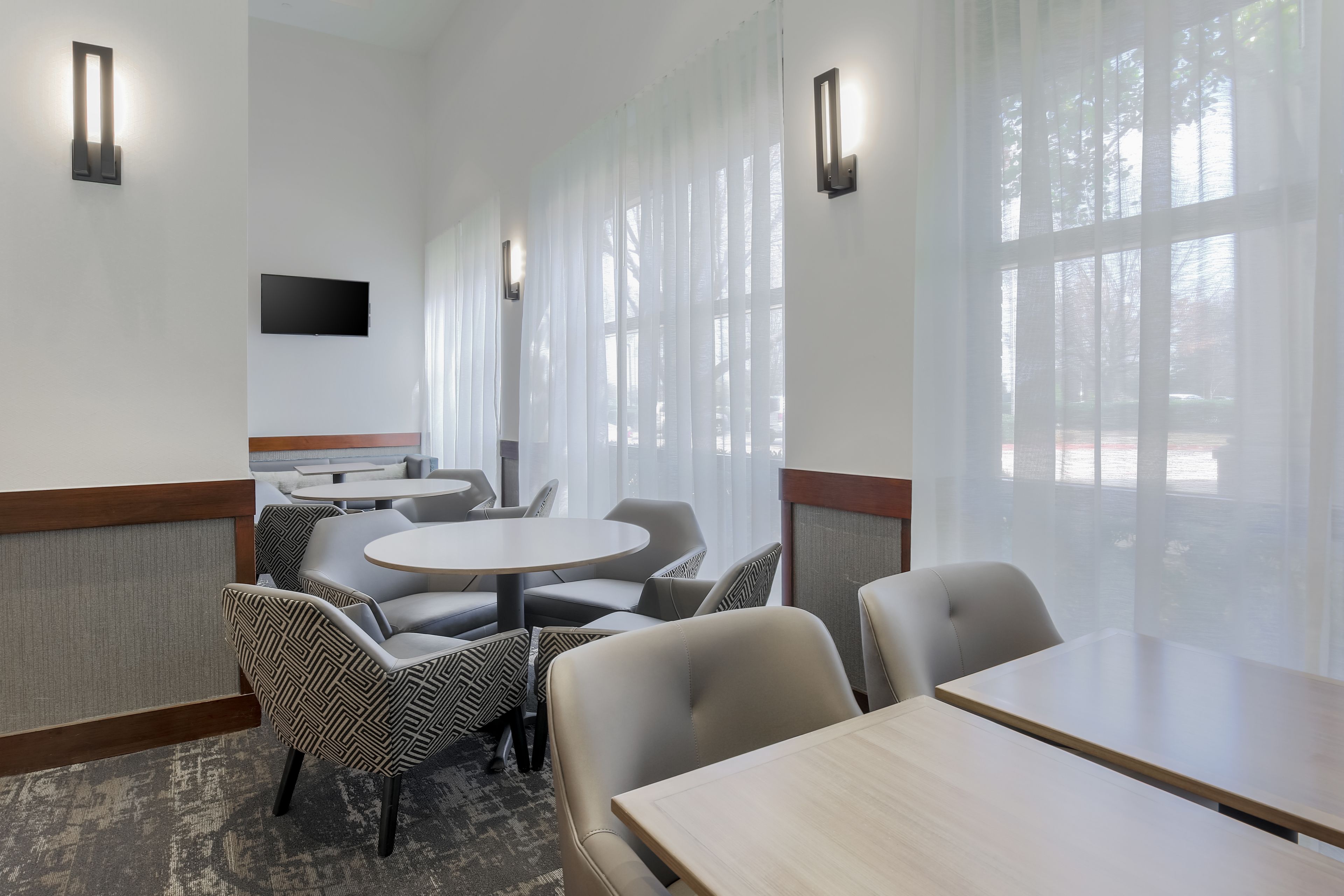 A hotel room with a large window, a flat-screen TV mounted on the wall, and a seating area with round tables and chairs.