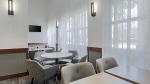 A hotel room with a large window, a flat-screen TV mounted on the wall, and a seating area with round tables and chairs.