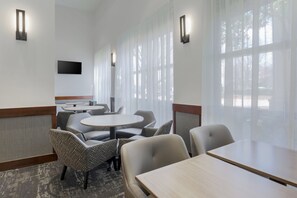 A hotel room with a large window, a flat-screen TV mounted on the wall, and a seating area with round tables and chairs.